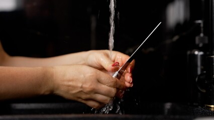 Hands cleaning a metal chef's knife under streaming water in a kitchen sink, ensuring hygiene for...