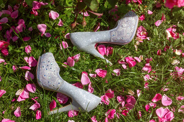 A pair of elegant silver high-heeled shoes, lying on the lush green grass, surrounded by a bright scattering of fresh rose petals.