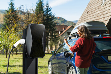 Woman traveler with smartphone next to charging electric car on backyard of country house