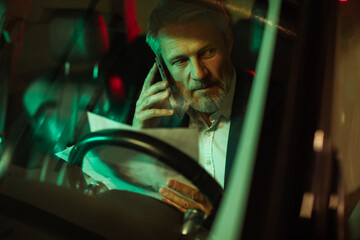 Mature man on phone reviewing papers in car at night, serious