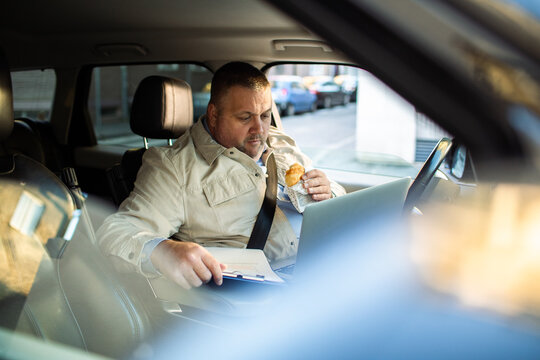 Adult man focused, working on laptop and eating breakfast in parked car