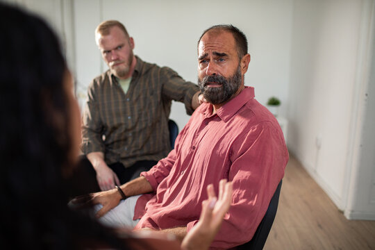 Adult man crying in group therapy, comforted by friend indoors