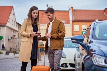 Man and woman with suitcase standing by car and looking at smartphone