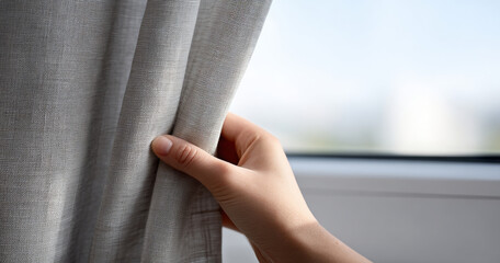 Close-up of hand gently pulling back light gray textured curtain near window with blurred outdoor background in soft natural light