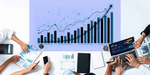 A diverse business team collaborates to analyze financial data displayed on charts and graphs, using laptops and notebooks in a modern workspace environment. Axiom