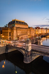 The aerial scene shows the National Theatre in Prague illuminated at dusk. The Smetana embankment...