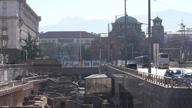 Handheld winter video in Sofia shows St Nedelya Cathedral, Serdica archaeological ruins, busy street with vehicles and pedestrians, and Sofia University buildings with distant mountain silhouettes
