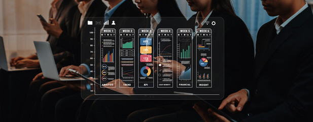 Business professionals collaborate and analyze data on laptops during a conference, with insightful graphs and charts displayed prominently on a digital screen. Trope