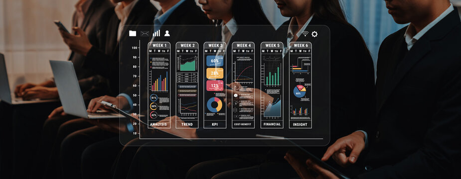 Business professionals collaborate and analyze data on laptops during a conference, with insightful graphs and charts displayed prominently on a digital screen. Trope - Powered by Adobe