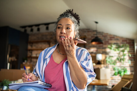 Mature woman focused, speaking on smartphone and taking notes at home