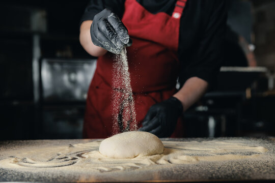 Baker pizzamaker preparing dough in pizzeria kitchen with red apron and gloves, dark light - Powered by Adobe