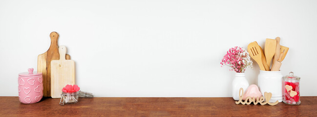 Valentines Day love and heart theme kitchen decor, bakeware and accessories on a wooden shelf against a white wall. Copy space.