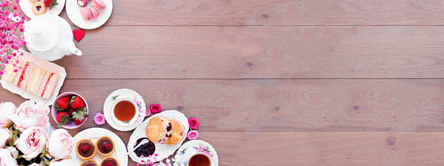 Valentines Day or love theme afternoon tea. Overhead view corner border against a wood banner background. Assortment of finger sandwiches, sweet desserts and pastries. Copy space.