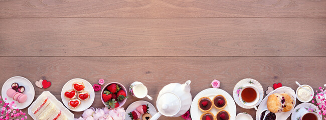 Valentines Day or love theme afternoon tea. Top down view bottom border against a wood banner background. Variety of finger sandwiches, sweet desserts and pastries. Copy space.