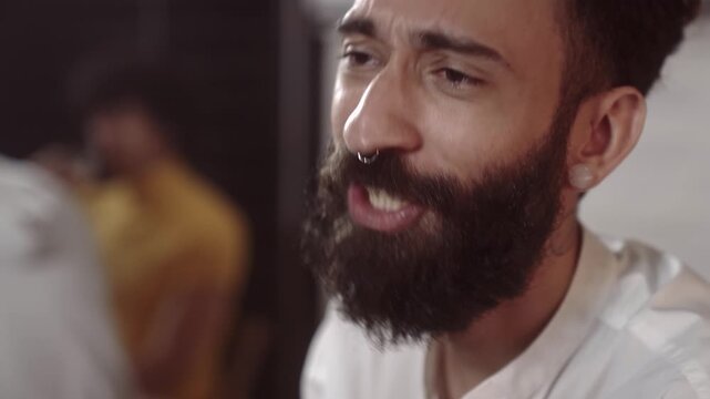 Close-up of a young stylish man with a thick beard, septum piercing, and ear gauges talking in a social indoor setting.