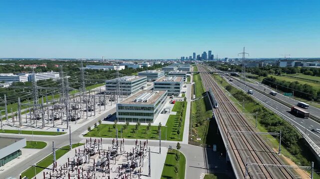 Aerial view of modern data center campus beside railway and highway, substation in foreground and city skyline beyond under clear blue sky, concept of urban cloud infrastructure