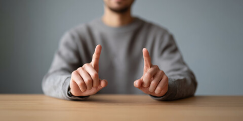 Man in gray sweater pointing with both index fingers forward sitting at wooden table with blurred background