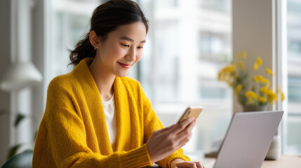 Young Asian woman in mustard yellow cardigan using smartphone while working on laptop at home desk near bright window cheerful remote work moment digital lifestyle multitasking