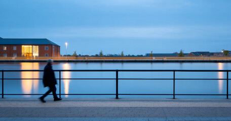 Silhouetted person walking along waterfront promenade during twilight with calm water and illuminated building reflections
