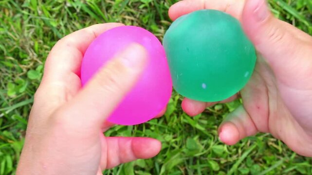 Close-up hands on green grass squeeze two colorful squishy water balloons, pressing them together, shifting grip and twisting, then releasing as the gel spheres rebound with glossy daylight.