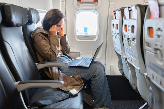 Frustrated adult caucasian woman holding her head while working on laptop inside airplane cabin. Stressful remote work and travel lifestyle concept