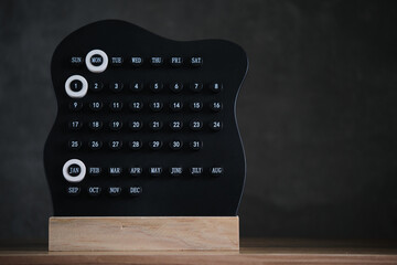 Modern black perpetual calendar on wooden base, with highlighted Monday and 1st January, standing against dark minimalist background.