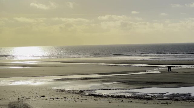 Two people walking on vast low tide beach between Quend and Fort-Mahon, France, with winding tidal pools and sunlit reflections under soft golden sunset sky
