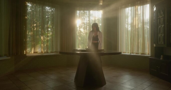 Girl enters sunlit living room, sits at round wooden table and spins book