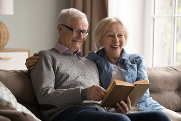 Cozy literary evening. Happy aged married couple enjoy calm family rest read paper book on comfy...