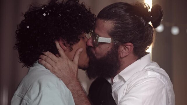 Romantic gay couple sharing an intimate kiss at a celebration with confetti in their hair. Diverse men expressing love and affection in a warm indoor setting.