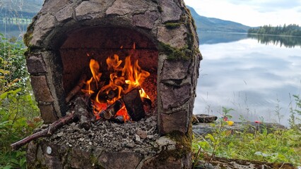 Campfire with stone oven next to lake during daytime in natural setting with trees and mountains in background