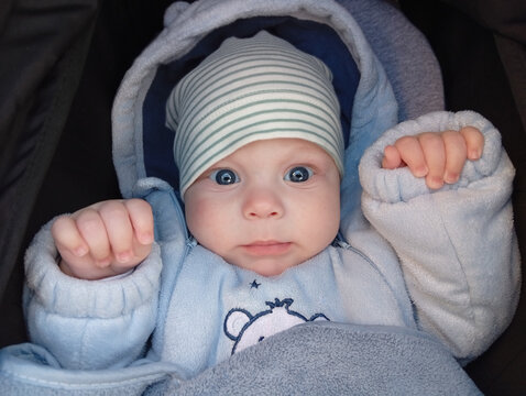 A baby with blue eyes wearing a light blue outfit and a striped hat is sitting in a stroller. The baby looks curious and is holding up their hands indoors during the day