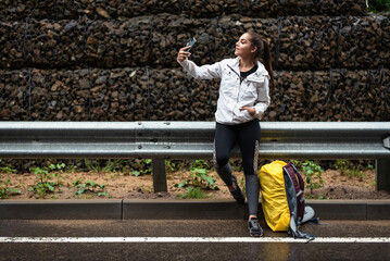 Young woman taking a selfie with her phone while on a hiking trip by the roadside