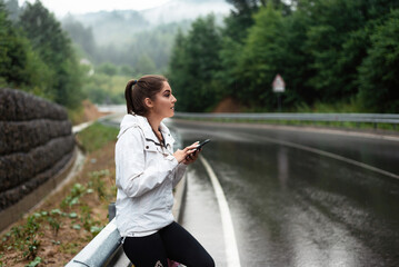 Young woman with smartphone on a wet road during rainy weather in the mountains