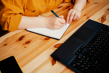 Woman writing notes in notebook at wooden desk with laptop nearby, cozy home office scene, focused work and creative planning lifestyle concept