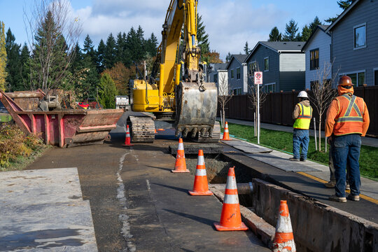 Excavator bucket attachment removing section of asphalt cut in road to dig a utilities trench, public infrastructure construction site, workman watching the project
