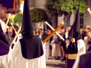 Nazarenes in white tunics and black hoods walking in a night procession with candles.