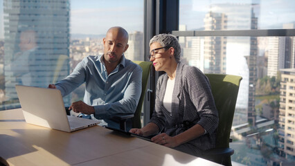 A male and female coworker analyze work on a laptop beside a large window with skyline views,...