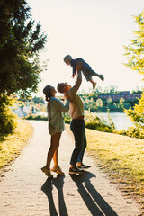 Fototapeta premium Family enjoying a sunny day by the riverside with a child being lifted in joy while walking on a path lined with greenery