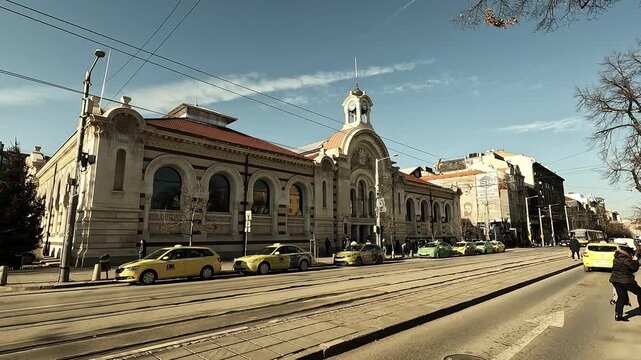 Handheld video of the central facade of Sofia Central Market Hall on a sunny winter day, with pedestrians passing by and light vehicle traffic in front, BG, EU, 30 Dec. 2025