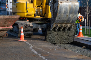 Excavator bucket attachment removing section of asphalt cut in road to dig a utilities trench, public infrastructure construction site, workman watching the project
