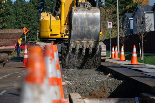 Excavator bucket attachment removing section of asphalt cut in road to dig a utilities trench, public infrastructure construction site, workman watching the project
