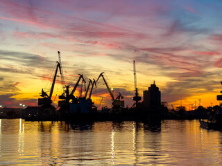 Obraz premium Sunset over a busy port with cranes reflecting in the water during evening hours