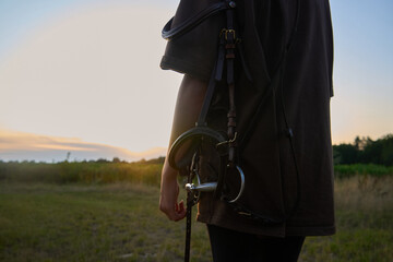 A girl with a horse harness and equipment stands in the summer at sunset and watches the sunset                               