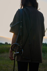 A girl with a horse harness and equipment stands in the summer at sunset and watches the sunset                               