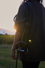 A girl with a horse harness and equipment stands in the summer at sunset and watches the sunset                               