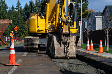 Excavator bucket attachment removing section of asphalt cut in road to dig a utilities trench, public infrastructure construction site, workman watching the project
