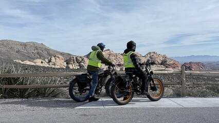 Obraz premium 🚲Back view of cyclists in yellow vests and helmets on e-bikes by a wooden fence. They are looking at the vast Red Rock Canyon desert landscape. Sunny day, travel and active lifestyle concept.