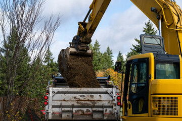 Excavator bucket attachment spilling dirt into a dump truck to haul away, public infrastructure construction site
