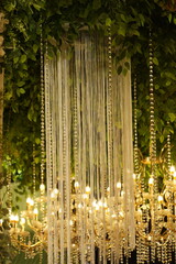 Luxurious interior wedding decor featuring a white string curtain and hanging crystal beads amidst lush artificial greenery, illuminated by warm golden chandeliers in a blurred background